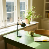DC LED Go Portable Table Lamp in dark green, with spherical shade on an oak strap and slim post, shown on a green desk near a sunlit window.