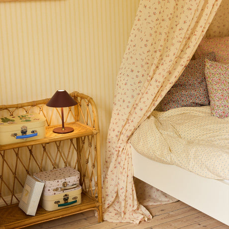 Hygge LED Rechargeable Outdoor Table Lamp in maroon with a wide cone shade and slim stem on a round base, shown on a wicker shelf in a bedroom setting.