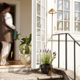 Hygge LED rechargeable outdoor floor lamp in antique brass with slim stem and cone shade, shown by a doorway with plants on a sunlit terrace.