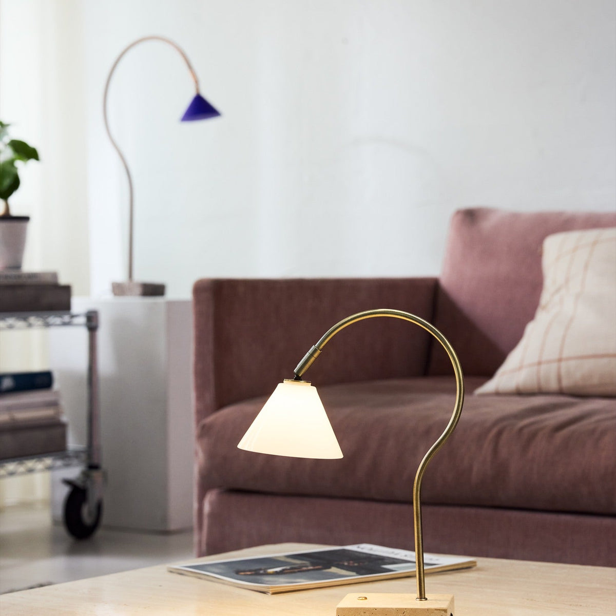 Question LED table lamp with a curved antique brass arm, conical opal glass shade, and travertine base, shown lit on a coffee table beside a mauve sofa.