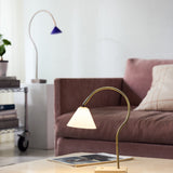 Question LED table lamp with a curved antique brass arm, conical opal glass shade, and travertine base, shown lit on a coffee table beside a mauve sofa.