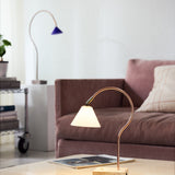 Question LED table lamp with a curved copper rod, conical opal glass shade, and antique brass fitting on a travertine base, shown lit on a coffee table in a living room.
