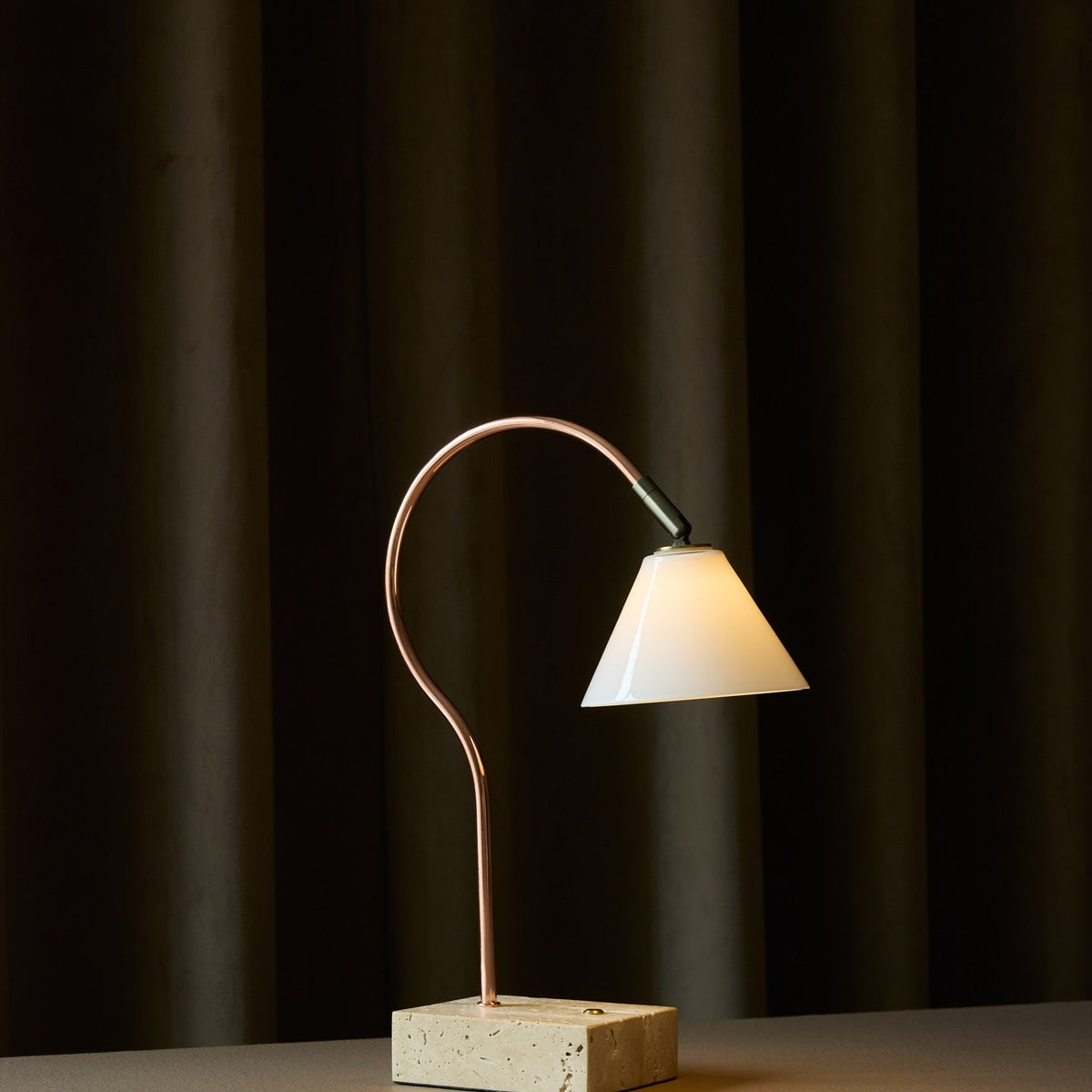 Question LED table lamp with a curved copper rod, conical opal glass shade on an antique brass knuckle, set on a travertine base, shown lit against a dark curtain backdrop.