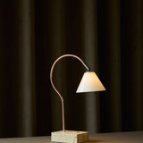 Question LED table lamp with a curved copper rod, conical opal glass shade on an antique brass knuckle, set on a travertine base, shown lit against a dark curtain backdrop.