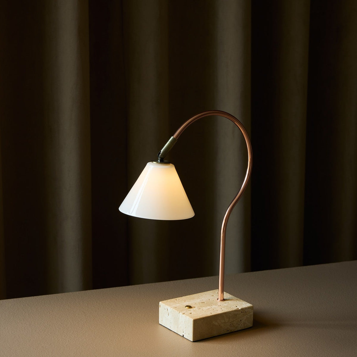 Question LED table lamp with a conical opal glass shade on a curved copper rod, set on a travertine base, glowing softly against dark curtains.