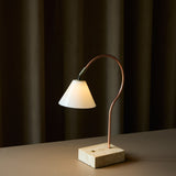 Question LED table lamp with a conical opal glass shade on a curved copper rod, set on a travertine base, glowing softly against dark curtains.