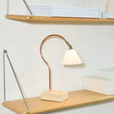 Question LED table lamp with a curved copper rod, opal glass conical shade, antique brass fitting, and travertine base, shown lit on a wooden shelf.