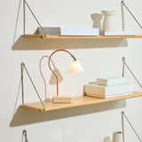 Question LED table lamp with curved copper rod, conical opal glass shade and antique brass fitting on a travertine base, shown lit on a wooden shelf.