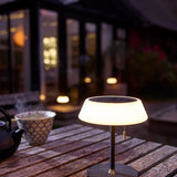 Solar LED Portable Outdoor Table Lamp in black, wide opal glass ring shade on a slim stem with brass pull toggle, glowing on an outdoor wooden table at dusk.