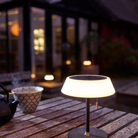 Solar LED Portable Outdoor Table Lamp in black, wide opal glass ring shade on a slim stem with brass pull toggle, glowing on an outdoor wooden table at dusk.