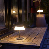 Solar LED Portable Outdoor Table Lamp in black, with a wide opal glass ring shade on a slim black stem and pull cord, shown lit on a wooden deck at night.
