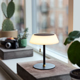 Solar LED Portable Outdoor Table Lamp in black, with a wide opal glass shade on a slim stem and brass pull toggle, shown lit on a windowsill beside plants.