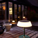 Solar LED Portable Outdoor Table Lamp in dark green, with an opal drum shade, black top panel, brass pull cord, and slim stem, glowing on an outdoor wooden table.