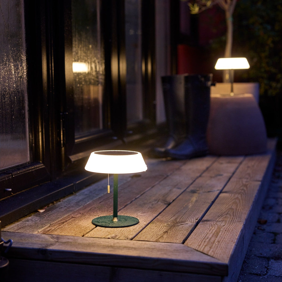 Solar LED Portable Outdoor Table Lamp in dark green, with wide opal drum shade, brass pull toggle, and slim stem, glowing on a wooden outdoor deck at night.