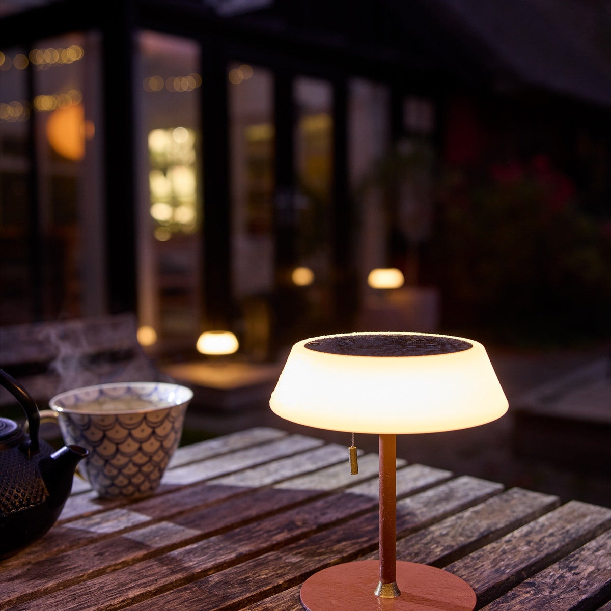 Solar LED Portable Outdoor Table Lamp in terracotta with a glowing opal drum shade and dark top panel, brass pull toggle, shown lit on a wooden table in an evening outdoor setting.