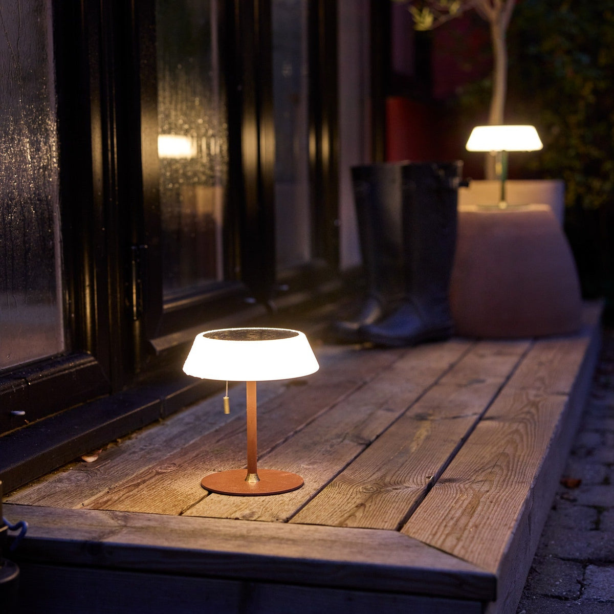 Solar LED Portable Outdoor Table Lamp in terracotta with wide opal drum shade, black top panel and brass pull cord, glowing on a wooden porch step at night.
