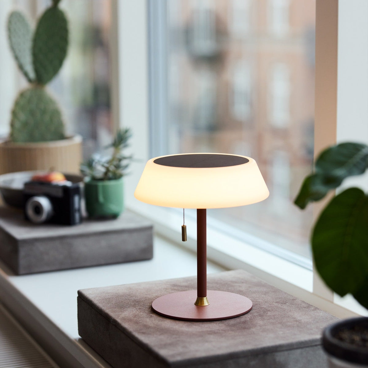 Solar LED Portable Outdoor Table Lamp in terracotta with opal drum shade, black top panel, and brass pull toggle, shown on a windowsill among plants.