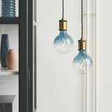 Two Colors Soft LED G125 3-Step Bulbs in blue gradient glass with S-shaped filaments, hung from black cables with brass fittings against a white wall.