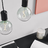 Colors Soft LED G125 3-Step Bulb in green, large round globe with S-shaped filament, shown lit in black pendant fittings above a workspace desk.