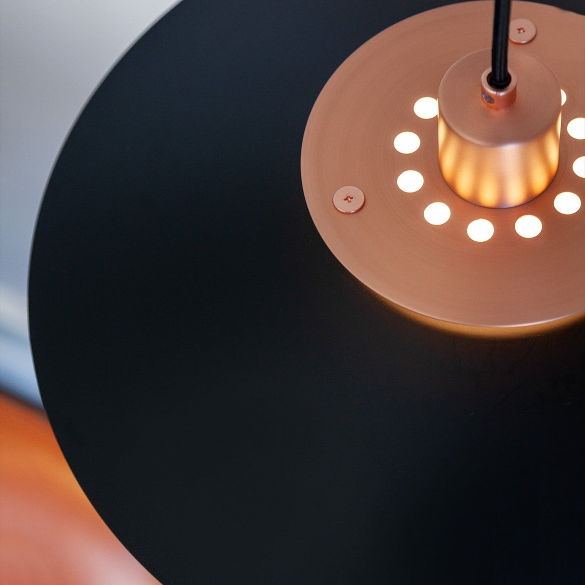 Close-up of the Rivoli Pendant Light 40cm in black and copper, showing the copper diffuser ring with circular cut-outs illuminated around a central cylinder beneath the flat black shade.
