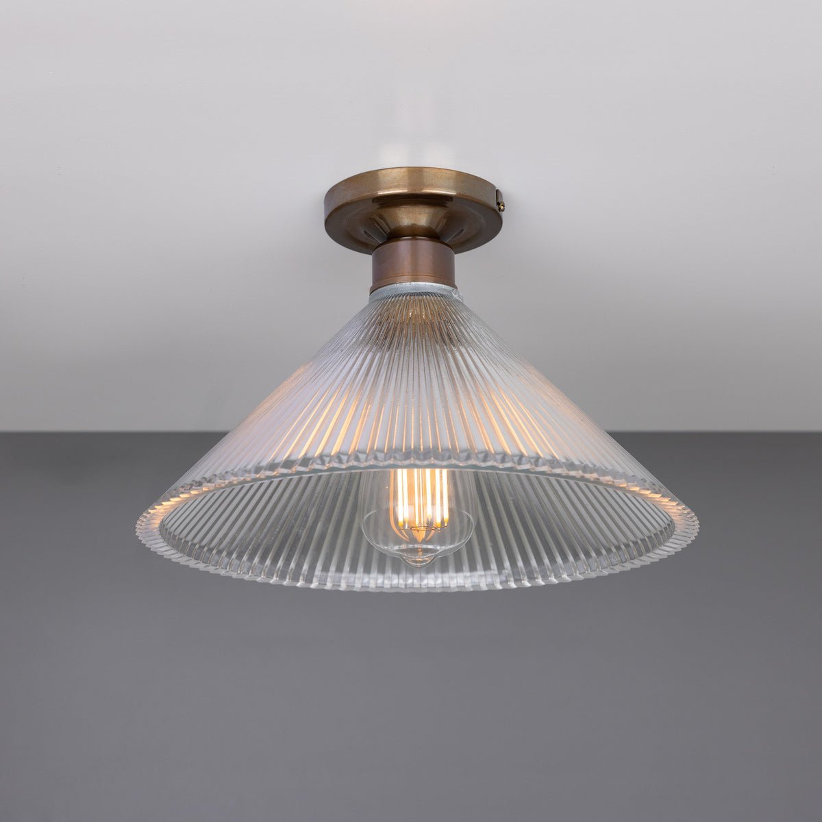 Hanoi Ribbed Glass Flush Ceiling Light with a cone-shaped clear ribbed glass shade and brass ceiling plate, shown lit with an exposed E27 bulb against a plain grey background.