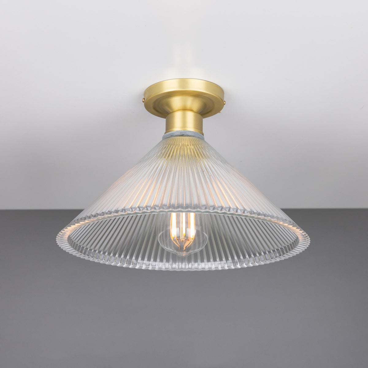 Hanoi Ribbed Glass Flush Ceiling Light with a cone-shaped prismatic glass shade and brass ceiling plate, shown with an exposed E27 bulb against a plain grey background.