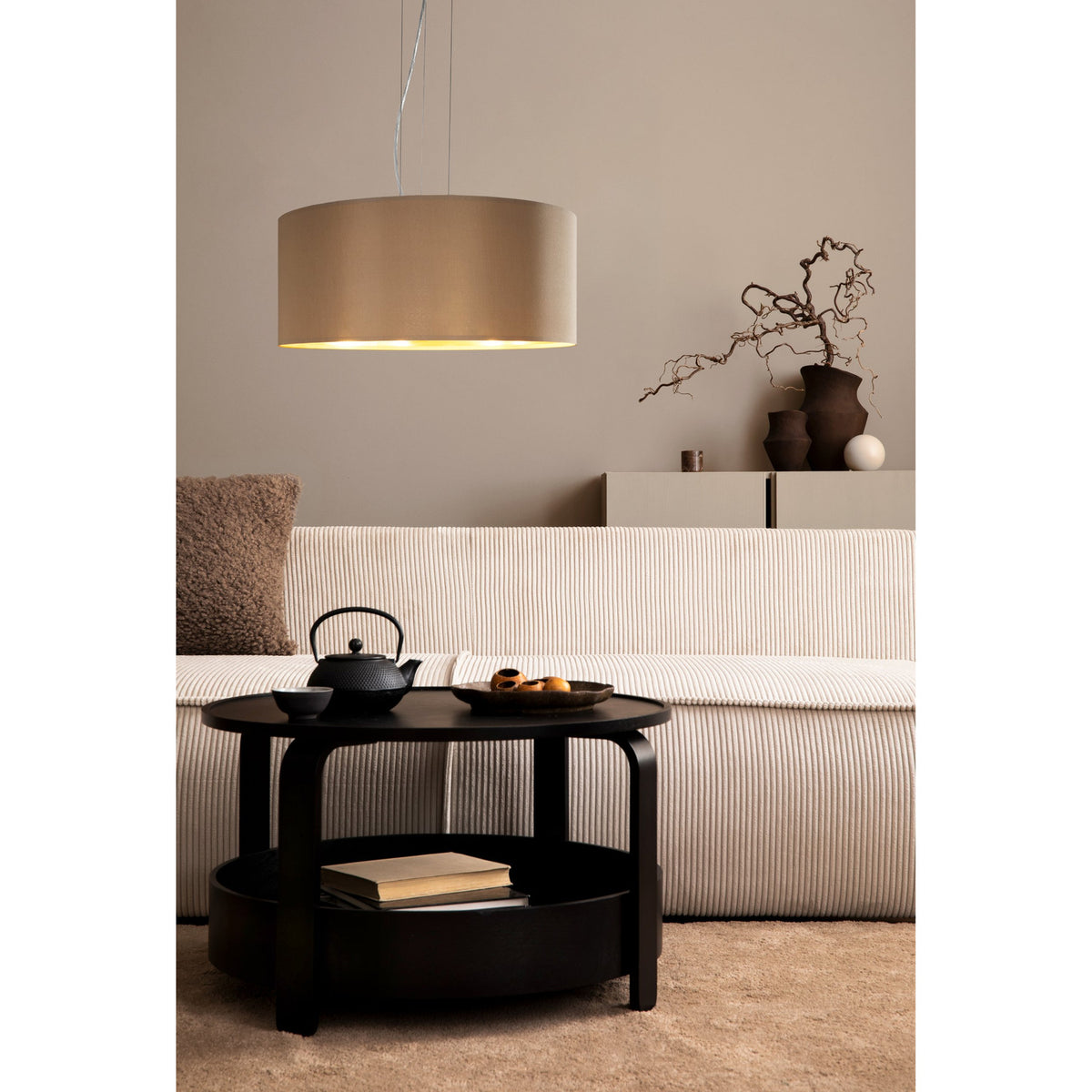 Hasanne 3 Light Pendant with wide taupe fabric drum shade, gold lining and satin nickel ceiling rose, hung above a coffee table in a living room.