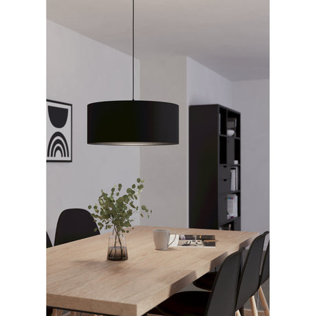 Hasanne Fabric Pendant Light in black, drum-shaped shade on a slim black cord, shown above a wooden dining table in a living space with dark shelving.
