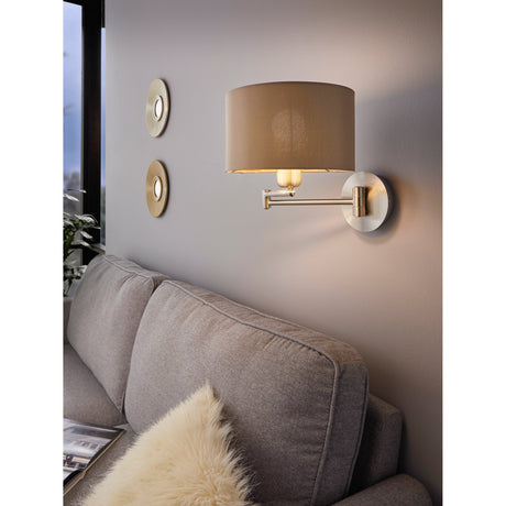 Hasanne Wall Light in satin nickel with a jointed swing-arm and taupe fabric drum shade, shown above a sofa in a living room setting.