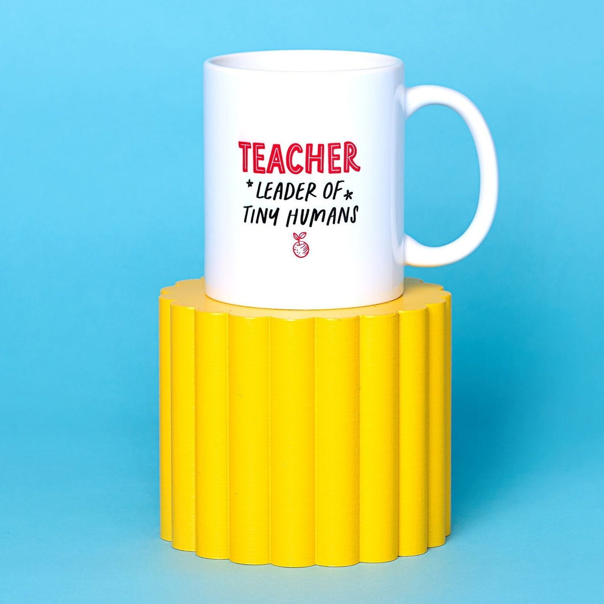 White stoneware mug with C-handle printed "TEACHER *LEADER OF* TINY HUMANS" and a red apple icon, displayed on a yellow stand against a blue background.