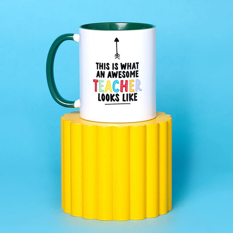 White stoneware mug with green interior and handle, printed with an upward arrow and This is what an awesome teacher looks like in black and rainbow lettering, on a yellow stand.