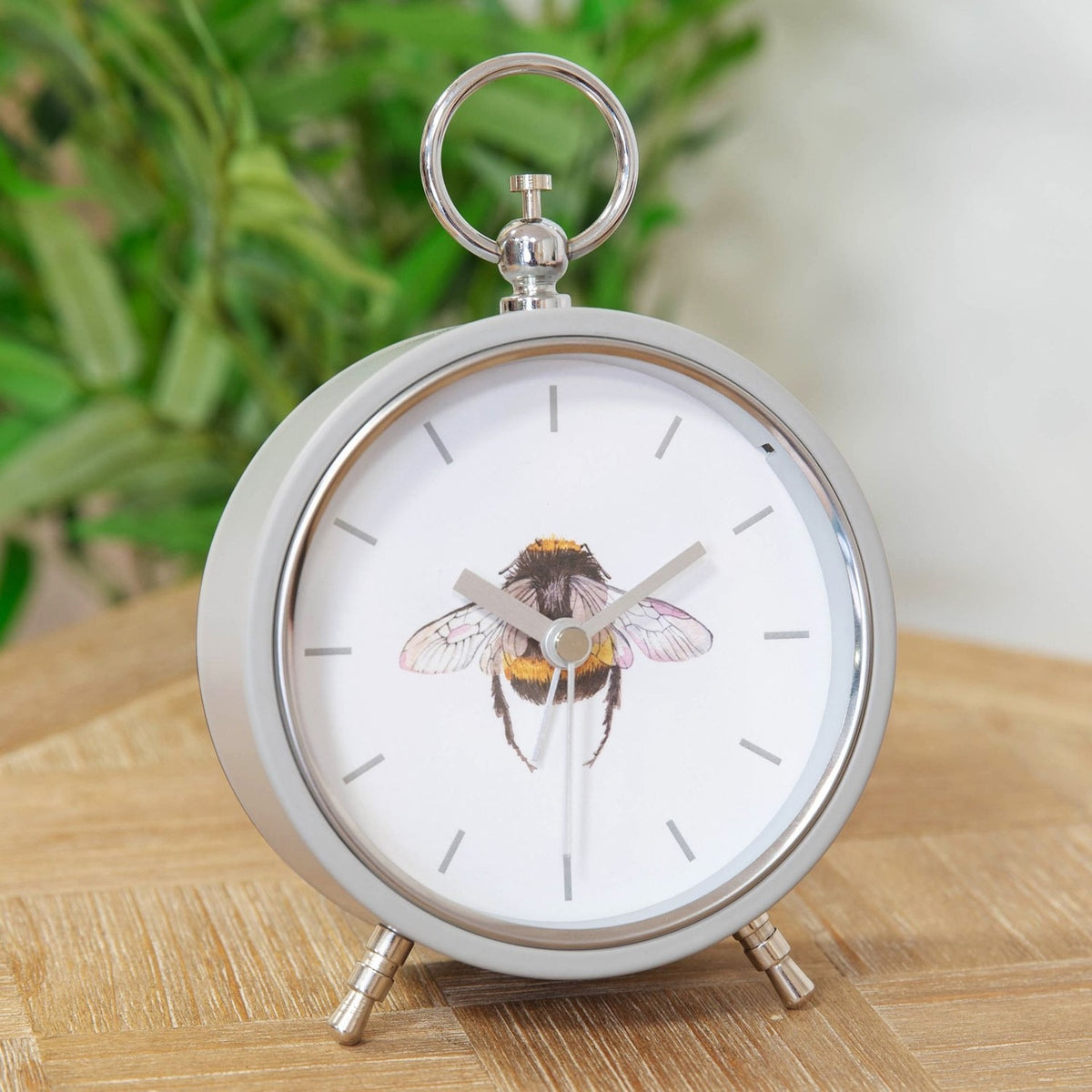 Round Bee Alarm Mantel Clock in grey metal with white dial, bumblebee illustration, baton markers, silver hands, top ring handle and angled feet, shown on a wooden surface.