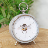 Round Bee Alarm Mantel Clock in grey metal with white dial, bumblebee illustration, baton markers, silver hands, top ring handle and angled feet, shown on a wooden surface.