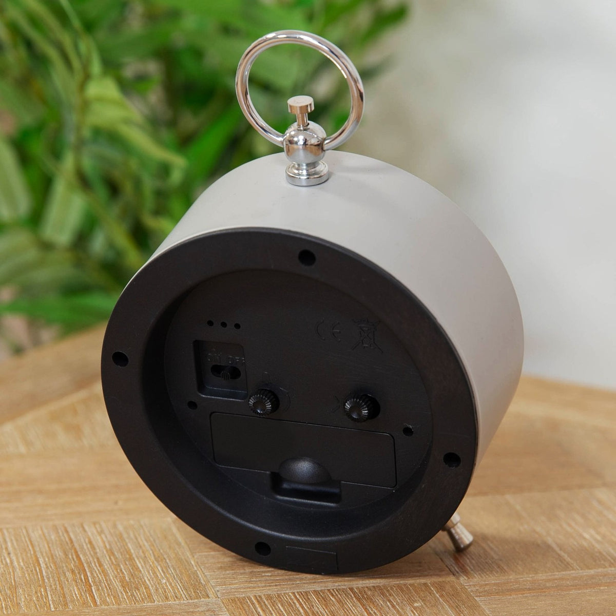 Rear view of the Bee Alarm Mantel Clock in grey metal, showing the chrome ring handle, snooze and time-set dials, and angled rear legs on a wooden surface.