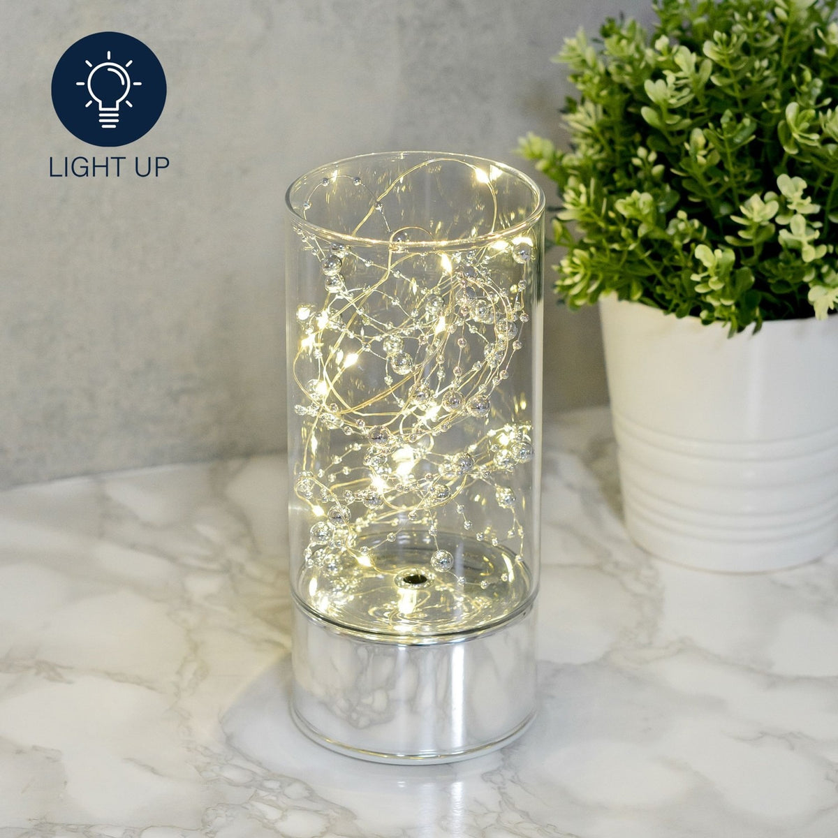 Cylindrical glass tube LED light-up decoration, 20cm, with warm fairy lights and silver beads on a mirrored silver base, shown on a marble surface beside a potted plant.