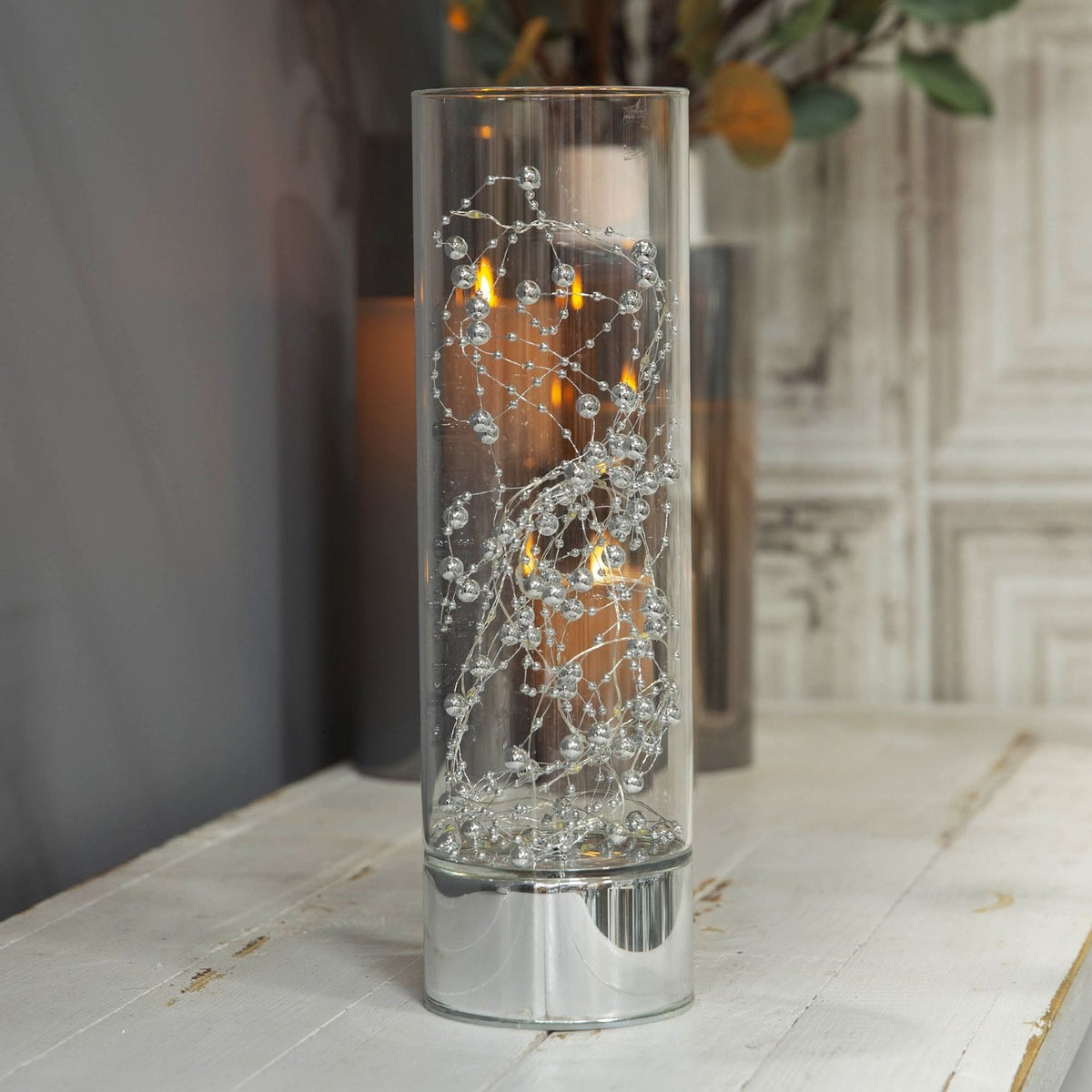 Beaded Glass LED Tube Light-Up Decoration, 30.5cm, with lit silver beads and wire inside a clear glass cylinder on a mirror-finish silver base, shown on a table in a living room setting.