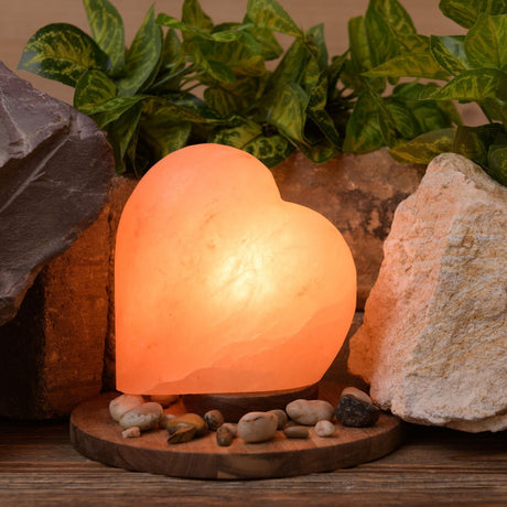 Heart salt lamp in pale pink with orange veining, lit on a round wooden base, surrounded by decorative stones and greenery.