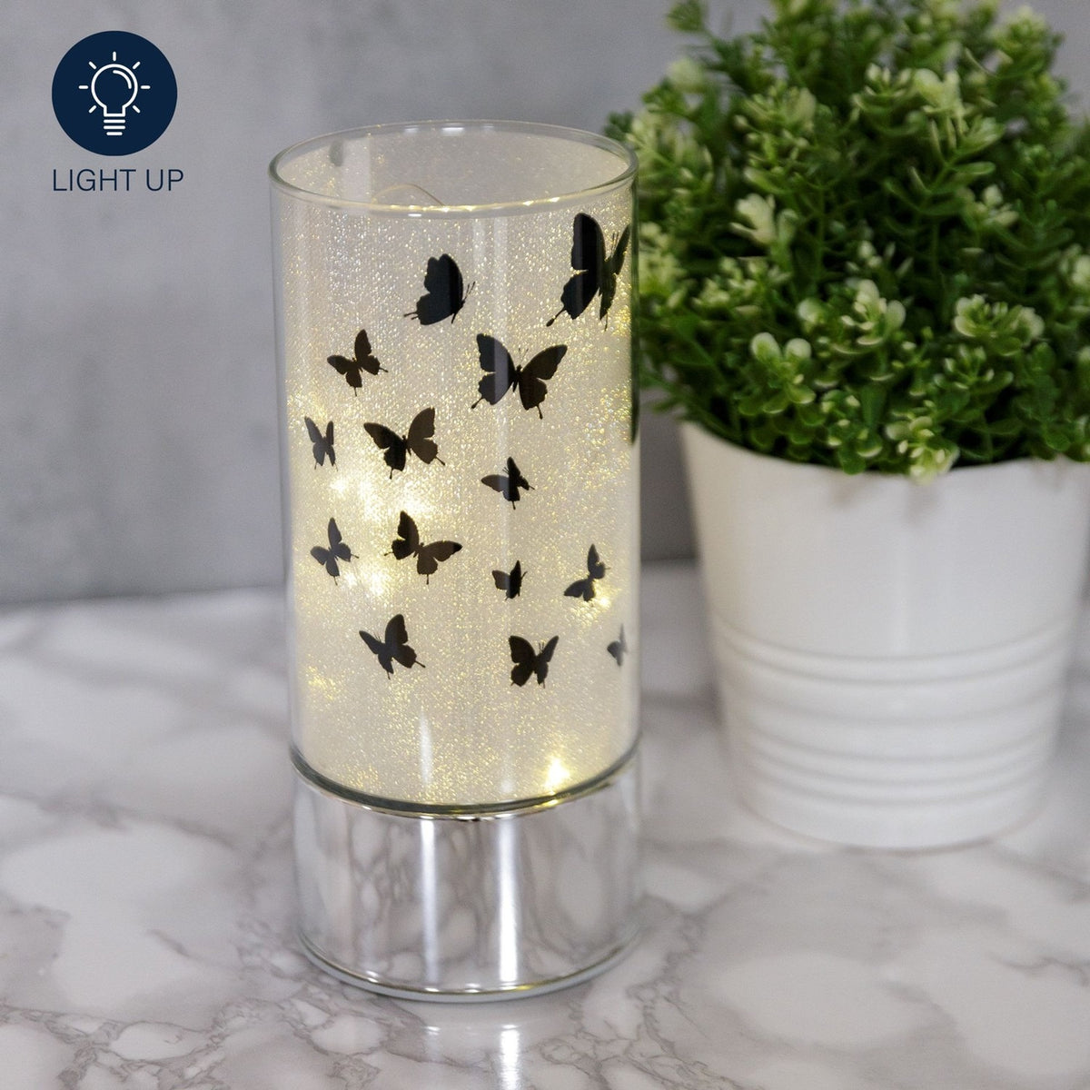 Butterfly Glass Tube LED Light-Up Decoration, 20.5cm, with black butterfly silhouettes on a glitter-lined glass cylinder and mirrored silver base, shown lit beside a potted plant.