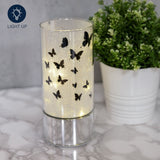 Butterfly Glass Tube LED Light-Up Decoration, 20.5cm, with black butterfly silhouettes on a glitter-lined glass cylinder and mirrored silver base, shown lit beside a potted plant.
