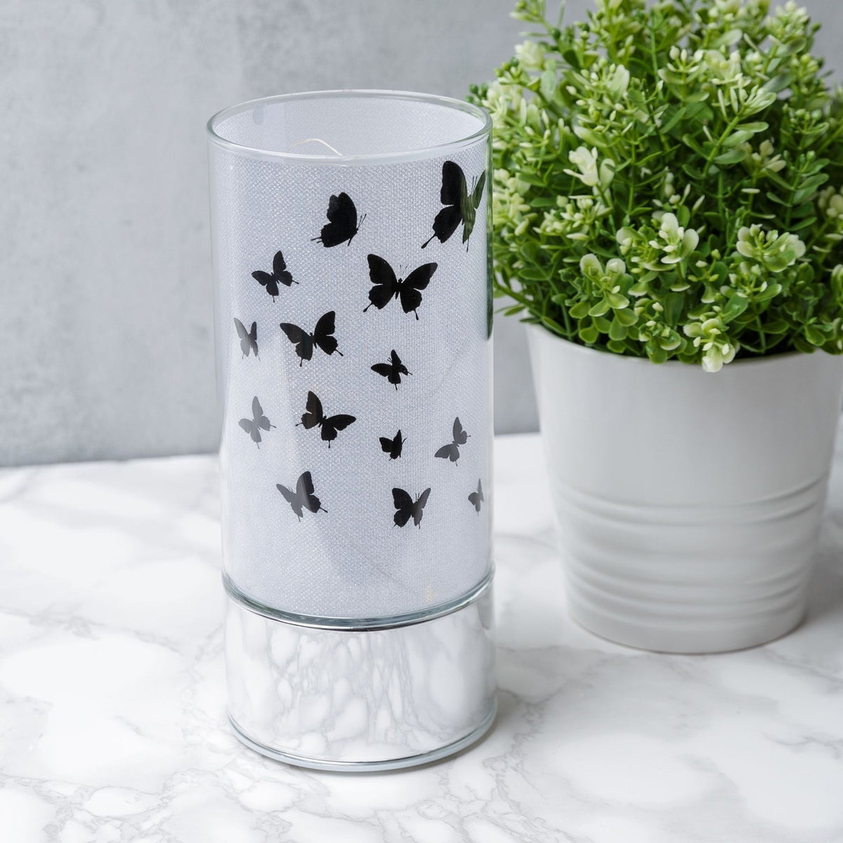 Butterfly Glass Tube LED Light-Up Decoration, 20.5cm, with black butterfly silhouettes and a mirrored silver base, displayed on marble beside a potted plant.