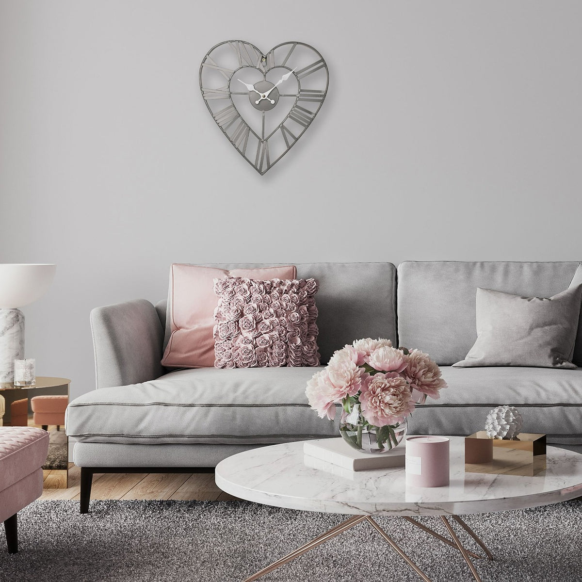Open frame heart-shaped wall clock in matt grey metal with Roman numerals, hung above a grey sofa with pink accent cushions in a living room setting.