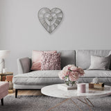 Open frame heart-shaped wall clock in matt grey metal with Roman numerals, hung above a grey sofa with pink accent cushions in a living room setting.