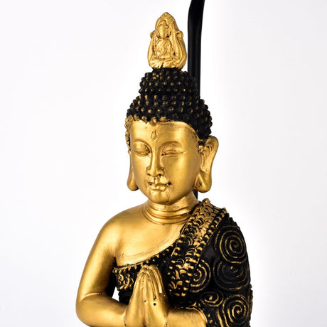 Close-up of the Praying Buddha Table Lamp, 46cm, showing the gold-faced figure with hands pressed together and a black robe patterned in raised gold swirls, against a plain white background.