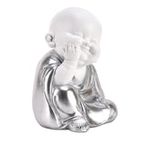 Buddha cherub figurine with white face and hands, sitting with hand covering eye and metallic silver draped robe, on a plain white background.
