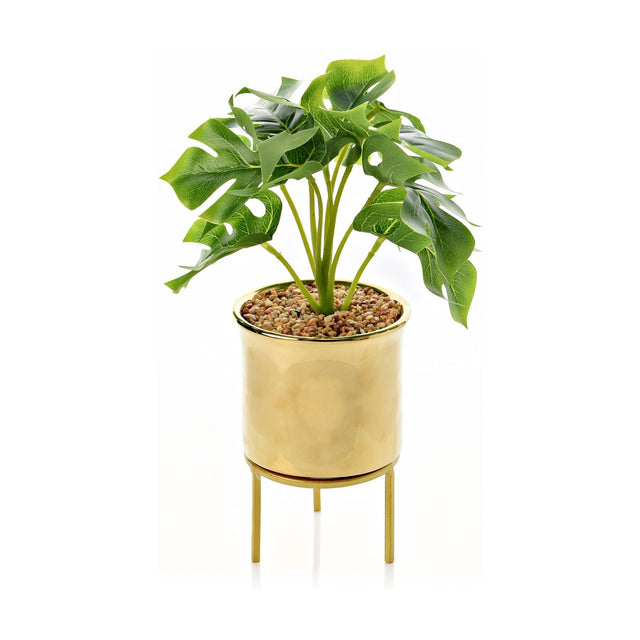 Footed gold iron planter with faux monstera foliage, glossy metallic cylindrical pot on a slim footed stand, 42cm tall, shown on a plain white background.
