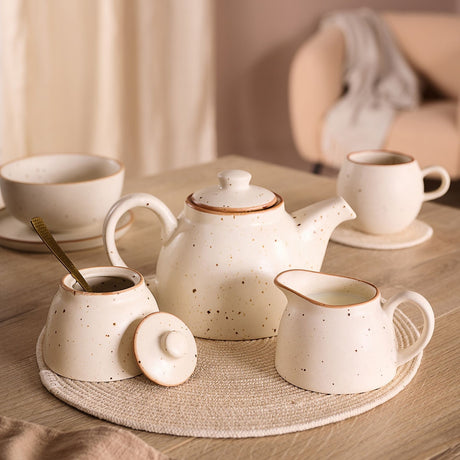 Speckled white stoneware tea set with brown rims, featuring a teapot with loop handle, lidded sugar pot with spoon, and milk pot, shown with cup and bowl on a wooden table.