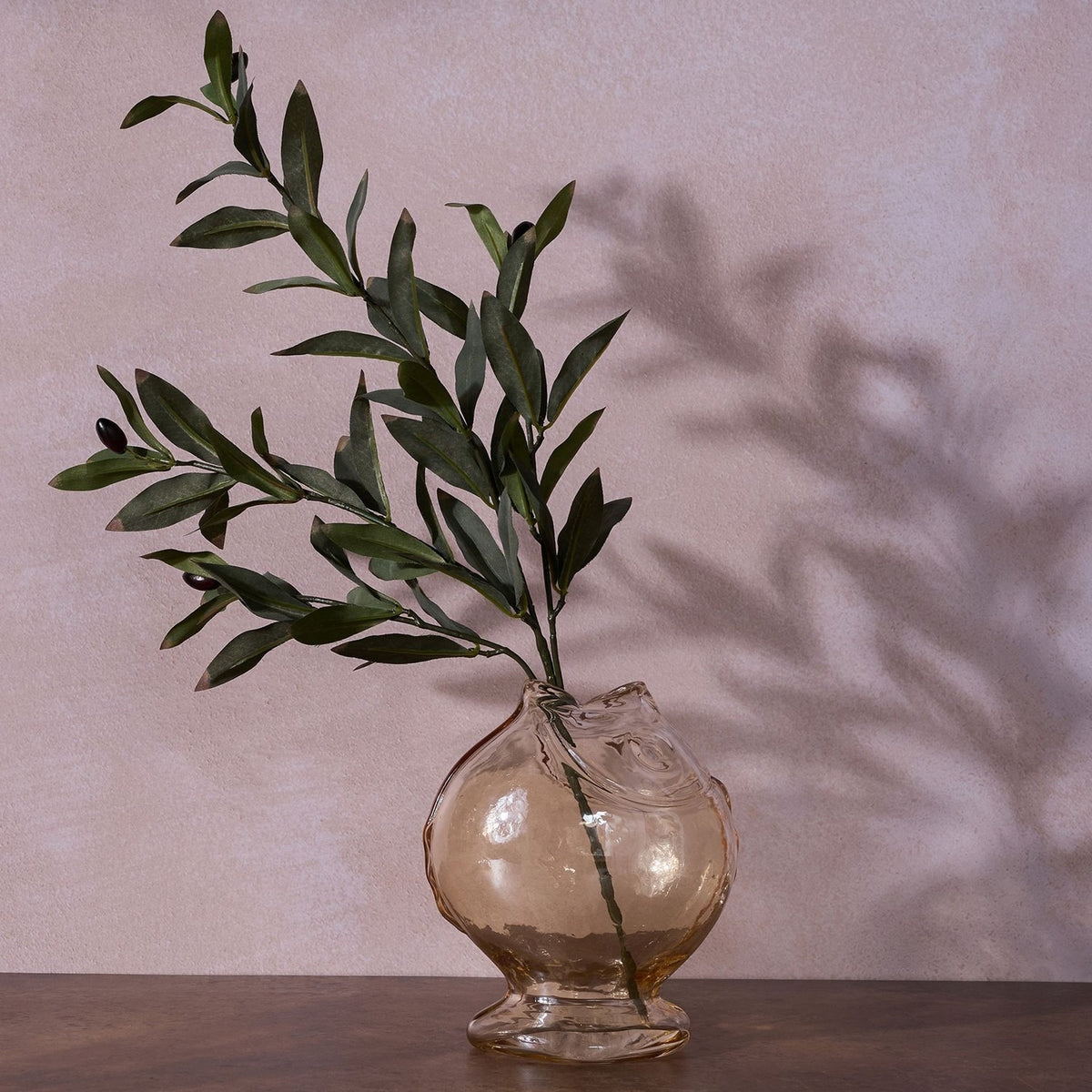 Round glass fish vase with footed base in clear glass with a pale amber tint, holding an olive branch, set against a pink wall with cast shadow.