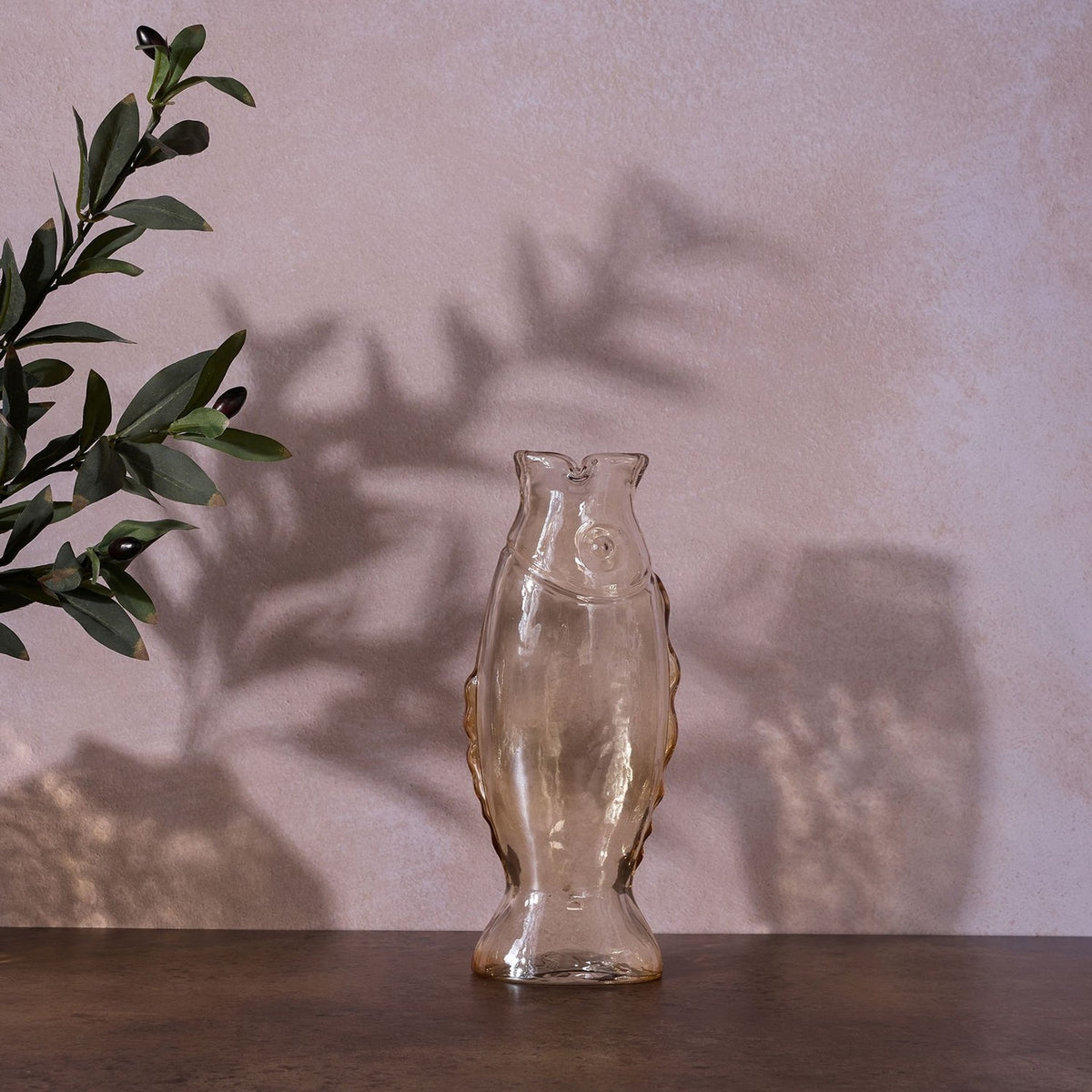 Tall Shiny Glass Fish Vase with Wavy Fins in clear glass, standing upright on its tail with a round eye and notched mouth opening, displayed against a pink wall beside an olive branch.