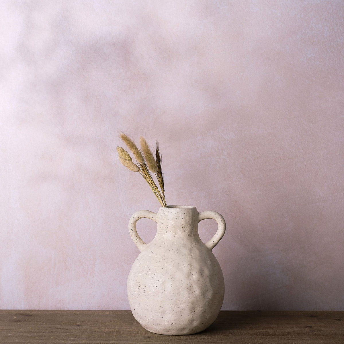 Round dimpled ceramic vase in cream with dark speckles, two looped handles, holding dried grasses, set on wood against a pink background.