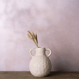 Round dimpled ceramic vase in cream with dark speckles, two looped handles, holding dried grasses, set on wood against a pink background.